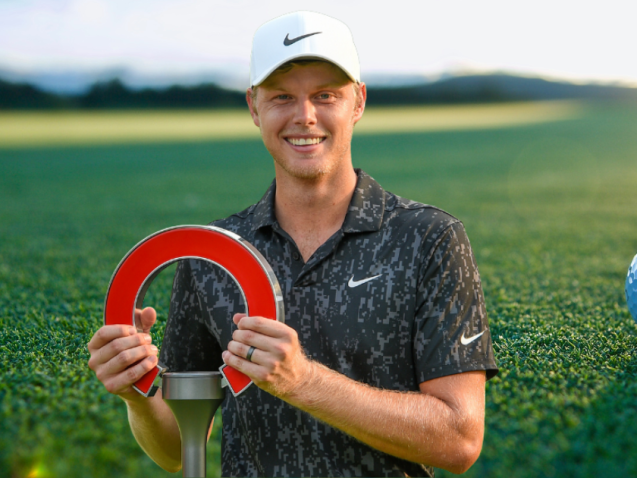 Golfer Cam Davis glimlachend met zijn trofee op de golfbaan, met een grote blauwe golfbal op de achtergrond.