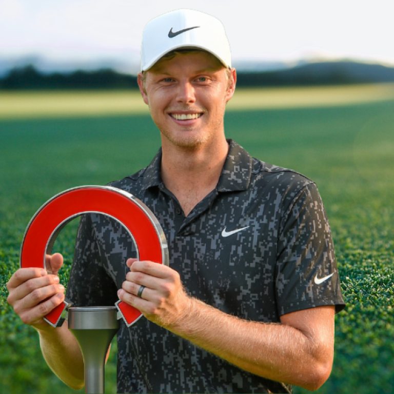 Golfer Cam Davis glimlachend met zijn trofee op de golfbaan, met een grote blauwe golfbal op de achtergrond.