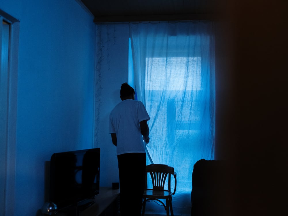Pleinvrees overwinnen met Hypnotherapie 2 Een persoon kijkt vanuit een donkere kamer door het gordijn naar buiten.