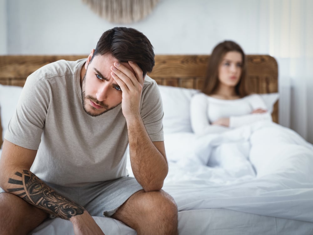 Een man zit op de rand van een bed met een vrouw op de achtergrond.